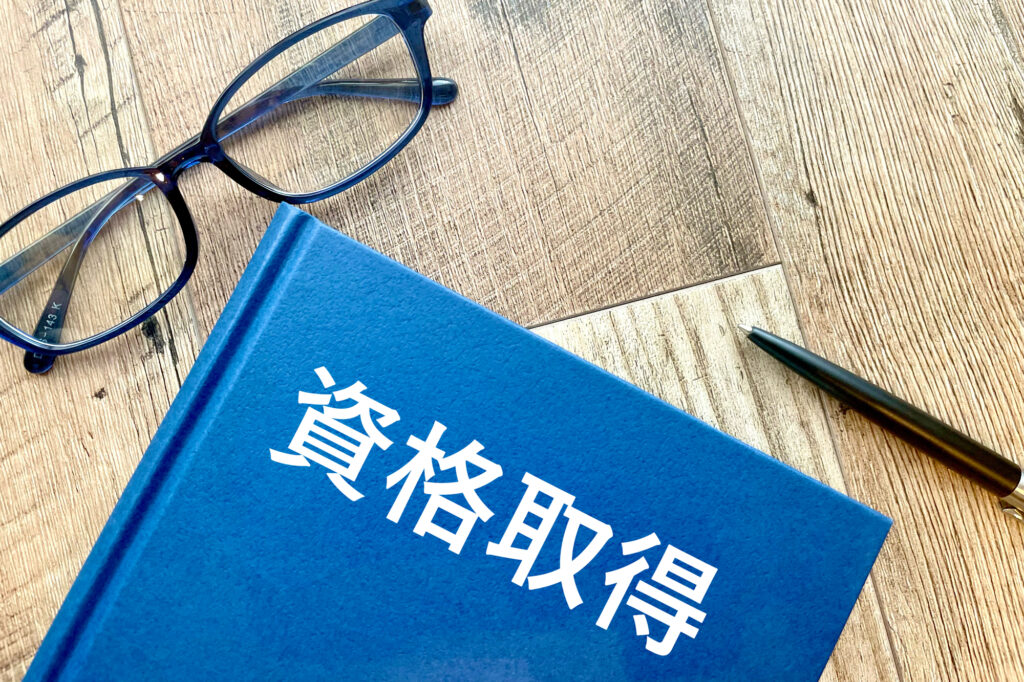 回路設計エンジニアにおすすめの資格のイメージ画像‐机の上に黒字で資格取得と書いてある青い参考書と黒縁メガネとボールペンが置かれている写真