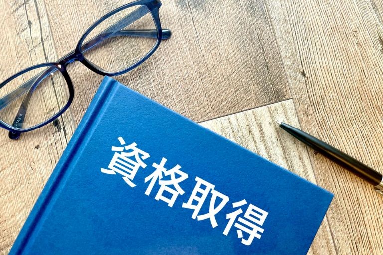 「設備管理の仕事に役立つおすすめの資格」のイメージ画像‐①白文字で資格取得と書かれた青い本と②黒フチの眼鏡と③ボールペンが机の上に置かれている画像